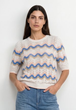 Donna con lunghi capelli scuri che indossa un maglione bianco lavorato a maglia con motivi a zigzag blu, rosa e beige e jeans blu, con le mani nelle tasche.
