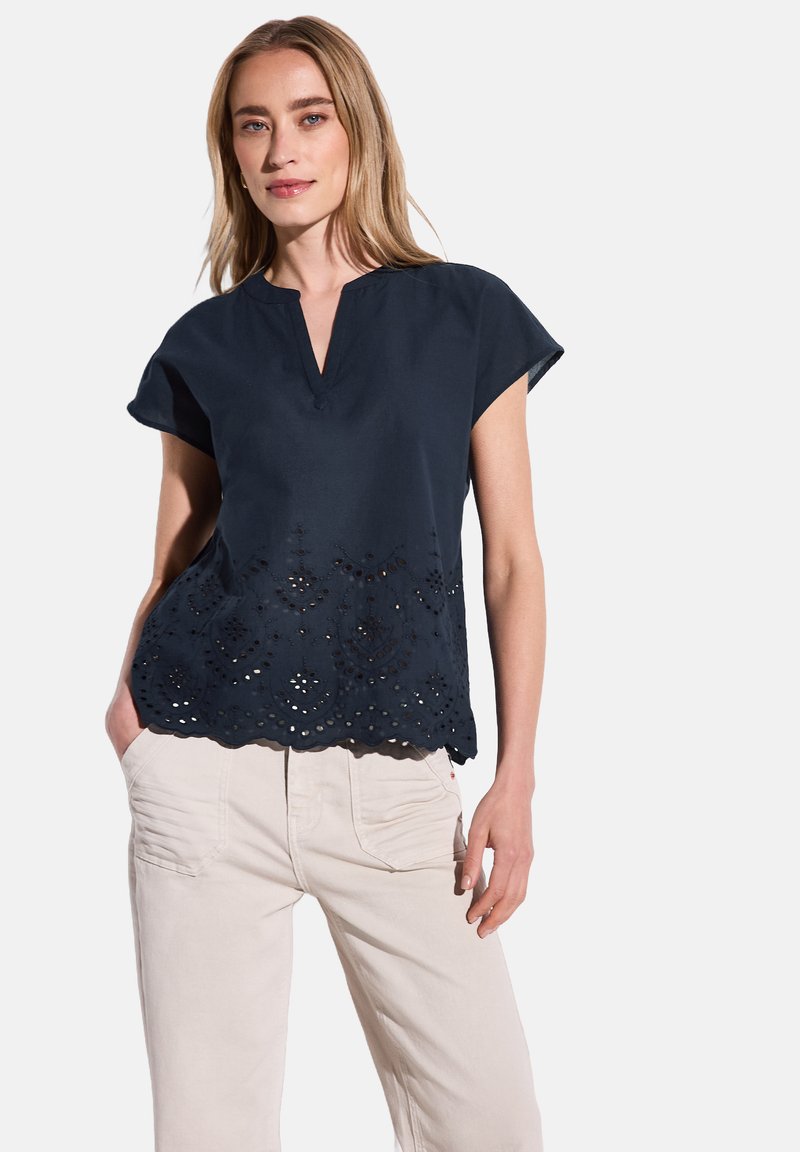 Camicia blu navy con maniche corte, scollatura a V e orlo scallopato con dettagli di motivi traforati. Abbinate a pantaloni di colore chiaro.