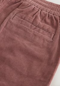 Primer plano de unos pantalones de pana rosa polvoriento que muestran una cintura elástica y un bolsillo trasero con abertura, con textura visible de canalé vertical.