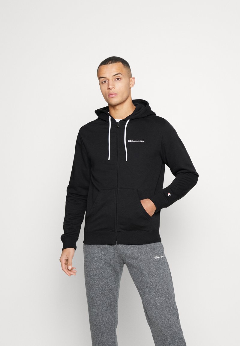 Felpa nera con zip, realizzata in misto cotone, con tasca frontale, stringhe bianche e un piccolo logo sul petto. Indossata con pantaloni felpati grigi.