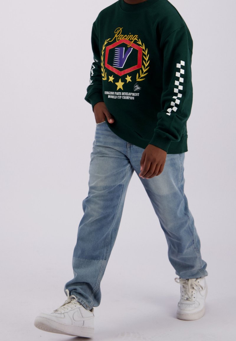 Personne portant un sweat-shirt de course vert foncé avec un logo graphique, un jean bleu clair et des baskets blanches, marchant avec une main dans la poche.