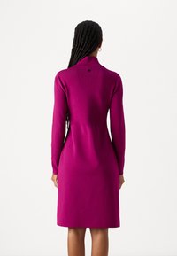 Figurbetontes Strickkleid in leuchtendem Pink, mit langen Ärmeln, hohem Kragen und mittellanger Saum; dezente Stickdetails auf der Rückseite.