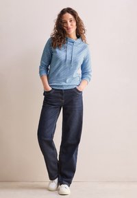 Ljusblå hoodie med snörning, bärs tillsammans med mörka jeans av denim. Jeansarna har synliga sömmar och framfickor; vita sneakers fulländar outfiten.