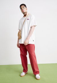 T-shirt en coton blanc avec logo, pantalon en denim rouge, sac bandoulière beige, chaussures de sport grises et rouges sur un sol vert.