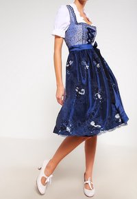 Robe dirndl bleu marine avec corsage en dentelle, manches bouffantes blanches, broderies florales et ruban en satin. Accompagnée de chaussures assorties.