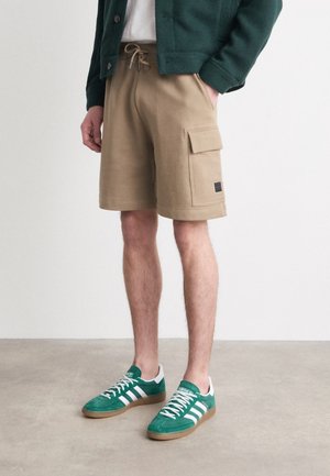 Mężczyzna ubrany w beżowe spodenki typu cargo z wiązanym paskiem w talii, ciemnozielony kardigan oraz zielone buty sportowe Adidas z białymi paskami i brązowymi podeszwami.