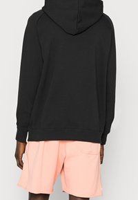 Felpa nera con cappuccio, dal fit morbido, maniche lunghe e polsini a coste, abbinata a pantaloni da allenamento rosa chiaro con tasca posteriore.