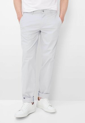 Mand iført lysegrå bukser med opkrogede ben, hvide sneakers og en hvid skjorte med hænderne i lommerne, stående mod en hvid baggrund.