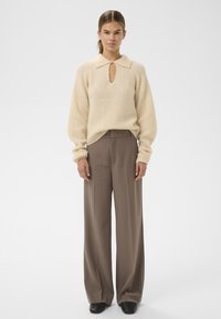 Kremkrāsas adīts džemperis ar atslēgas cauruma dekoltē, kombinēts ar augsto jostas līniju brūniem plašajiem biksēm un melnām kurpēm.