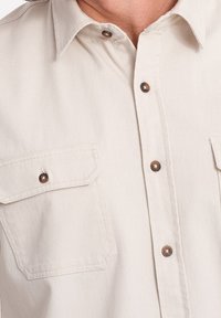 Camisa beige claro abotonada, hecha de algodón, con dos bolsillos en el pecho, botones de color cobre y un diseño de cuello clásico.