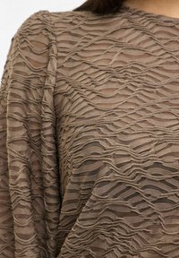 Haut marron texturé avec un tissu superposé ondulé présentant des lignes horizontales, créant un effet ondulé. Matière transparente laissant une certaine visibilité en dessous.
