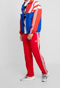 Trackjacka i blått, rött och vitt med stripdetaljer; matchad med röda träningbyxor med tre vita ränder; grå sneakers.