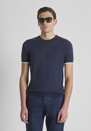 Uomo che indossa una maglietta a maniche corte a maglia blu scuro, pantaloni blu navy abbinati e occhiali da sole neri, in piedi contro uno sfondo neutro.