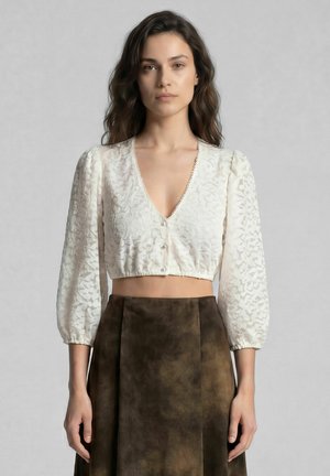 Femme aux cheveux mi-longs ondulés porte un chemisier court blanc texturé avec des manches bouffantes et une jupe marron taille haute.