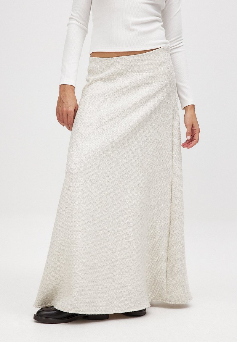 Langer weißer strukturierter Rock im Wickeldesign, aus einem weichen Stoff mit einer fließenden Silhouette, kombiniert mit dunklen flachen Schuhen.