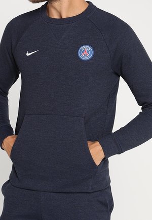 Granatowa bluza z kapturem z logo Paris Saint-Germain i białym znakiem Nike swoosh, noszona przez osobę z rękami w przedniej kieszeni.