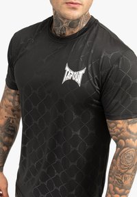 Melns sporta krekls ar vispārēju smalku rakstu un izteiktu balto "TAPOUT" logo uz krūtīm. Īsas piedurknes, gluda tekstūra.