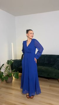 Mulher com vestido azul royal fluido de mangas compridas, a sorrir, com as mãos na anca, perto de um sofá verde e de uma planta em vaso no interior.