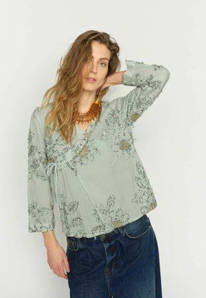 Femeie cu păr lung și ondulat, purtând o bluză florală verde deschis și blugi albastru închis, pozând cu un braț ridicat în spatele capului.