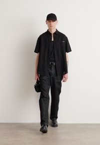 Camisa negra de botones de manga corta, camiseta negra, pantalones oscuros con estampado, gorra de béisbol negra, sosteniendo una cámara, de pie sobre un fondo neutro.
