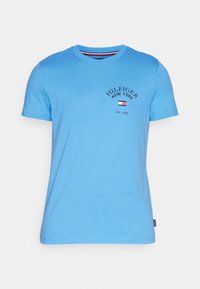 Světle modré tričko s krátkým rukávem a kulatým výstřihem s textem "Hilfiger New York Est. 1985" a malou vlajkou na levé části hrudi.