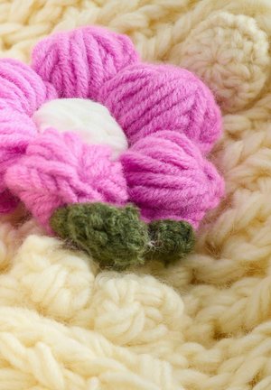 Rozā dzijas zieds ar baltu centru un zaļām lapām, novietots uz mīksta, teksturēta bēša adījuma fona.