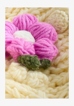 Rožinis siūlų žiedas su baltu centru ir žaliais lapeliais, padėtas ant minkšto, tekstūruoto smėlio spalvos megzto fono.