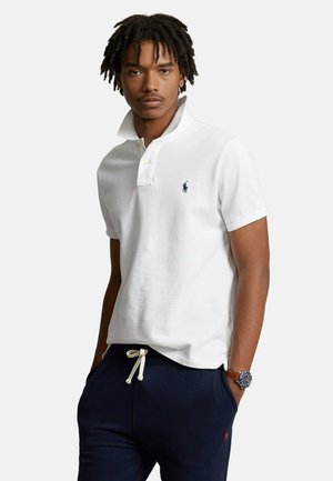 Giovane uomo con capelli scuri che indossa una polo bianca con colletto e pantaloni blu navy con coulisse, in piedi con le mani nelle tasche.