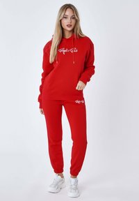 Felpa rossa con cappuccio e pantaloni coordinati in tessuto morbido, con tasca frontale e logo sul petto e sulla gamba. Sneakers bianche completano il look.