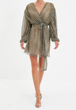 Kobieta ubrana w metaliczną, plisowaną sukienkę kopertową z długimi bufkami i wiązanym paskiem w talii, w połączeniu z sandałami na wysokim obcasie z odkrytymi palcami.