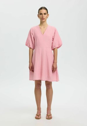 Kobieta stojąca, ubrana w różową sukienkę o fakturze do kolan z dekoltem w serek i bufkami na krótkich rękawach, w połączeniu z brązowymi sandałami z odkrytymi palcami.