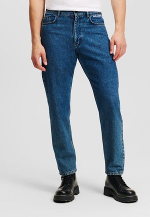 Jean en denim bleu avec une coupe droite, design à cinq poches et coutures contrastées. Porté avec des bottes noires à lacets.