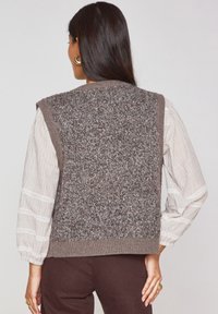 Gilet lavorato a maglia marrone con schiena testurizzata, bordo a coste e design senza maniche, abbinato a una blusa leggera a righe con maniche a palloncino.