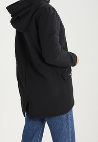 Czarna kurtka z kapturem o luźnym kroju, z kieszenią typu kangur, złotymi detalami oraz dłuższym tyłem. Gładka tekstura.