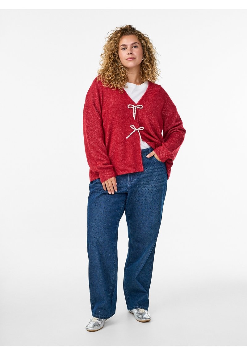 Vrouw met krullend haar die een rood vest met witte strikken, een wit overhemd, blauwe spijkerbroek en zilveren schoenen draagt, staand met één hand in de zak.