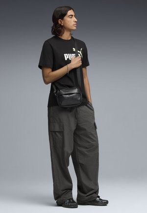 Joven con camiseta negra de Puma, pantalones cargo oscuros y holgados, zapatillas deportivas y bolso cruzado negro, posando frente a un fondo gris.