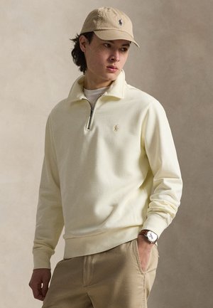 Νέος άνδρας που φοράει μπεζ καπέλο, κρεμ φούτερ με φερμουάρ στο λαιμό, χακί παντελόνι και ρολόι χειρός, στέκεται μπροστά σε μονόχρωμο μπεζ φόντο.