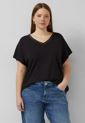 Junge Frau mit glattem Haar, die Creolen trägt, ein schwarzes V-Ausschnitt-Top mit Spitzenbesatz und blaue Jeans, steht mit einer Hand in der Tasche.