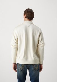 Sweatshirt color crema con colletto e polsini a coste, caratterizzato da un'accentuazione a forma di V sul retro, abbinato a jeans blu scuro.