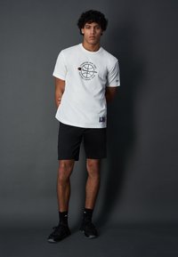 T-shirt bianca in cotone con design del logo nero, abbinata a pantaloni corti sportivi neri. Modello con scarpe e calze nere. Sfondo a tinta unita.