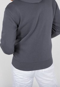 Felpa grigia scura con una texture morbida, polsi e vita a costine; presenta un cappuccio e una vestibilità rilassata. Indossata con pantaloni bianchi.