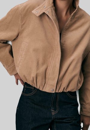 Beige korte ribfluwelen jas met knoop- en ritsluiting, elastische taille, gedragen over donkerblauwe spijkerbroek met contrasterend stiksel.