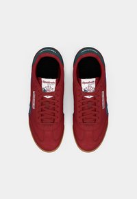 Červené semišové tenisky s modrými akcentmi, vybavené gumovou podrážkou, okrúlym prstom a dizajnom na šnurovanie. Obsahujú branding Reebok na jazyku.