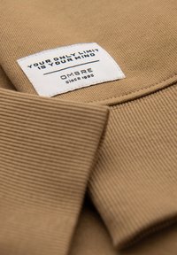 Gros plan sur un tissu côtelé beige avec une étiquette rectangulaire blanche portant l'inscription "VOTRE SEULE LIMITE EST VOTRE ESPRIT" et "OMBRE depuis 1985".