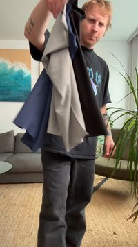 Četri salocīti krekli melnā, pelēkā, tumši zila un gaiši pelēkā krāsā, ko tur kāda persona ikdienas apģērbā, fonā redzama augu un dīvāns.