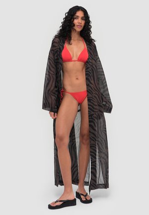 Jonge vrouw draagt een rode bikini en een doorschijnende zwarte lange cover-up met zebraprint, staand met zwarte plateau flip-flops tegen een neutrale achtergrond.