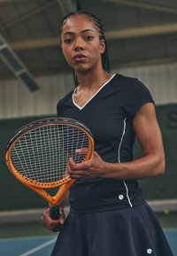 Μαύρη αθλητική μπλούζα με λευκές λεπτομέρειες και κοντά μανίκια, σε συνδυασμό με σκούρα φούστα. Ο παίχτης κρατά ένα πορτοκαλί και μαύρο τένις ρακέτα.