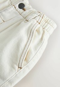 White denim pants feature elastic waistband, side pocket, and contrasting brown stitching on seams and openings. Round black button accents.