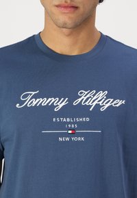 Sötétkék pamutt póló, fehér hímzett szöveggel: "Tommy Hilfiger", "Established 1985" és "New York", egy kis logó részlettel.