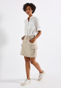 Beige knälång kjol med cargofickor, en enda knapp och dragkedjestängning, tillsammans med en vit linneskjorta och sneakers.